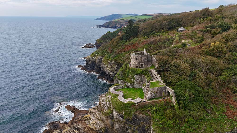 Aerial view of a coastal cliff with a historic stone tower and grassy area overlooking the ocean, surrounded by rugged rocks and lush foliage.