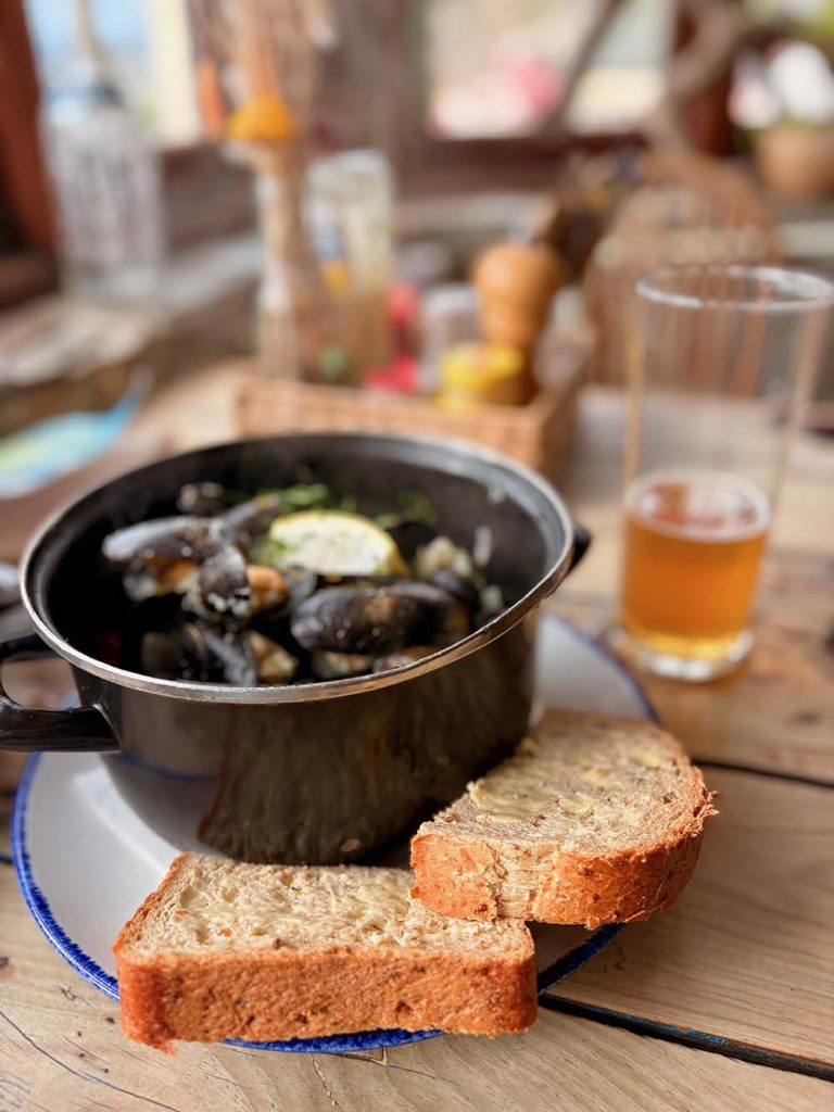 A bowl of cooked mussels served with slices of brown bread on a wooden table, accompanied by a glass of beer in the background.
