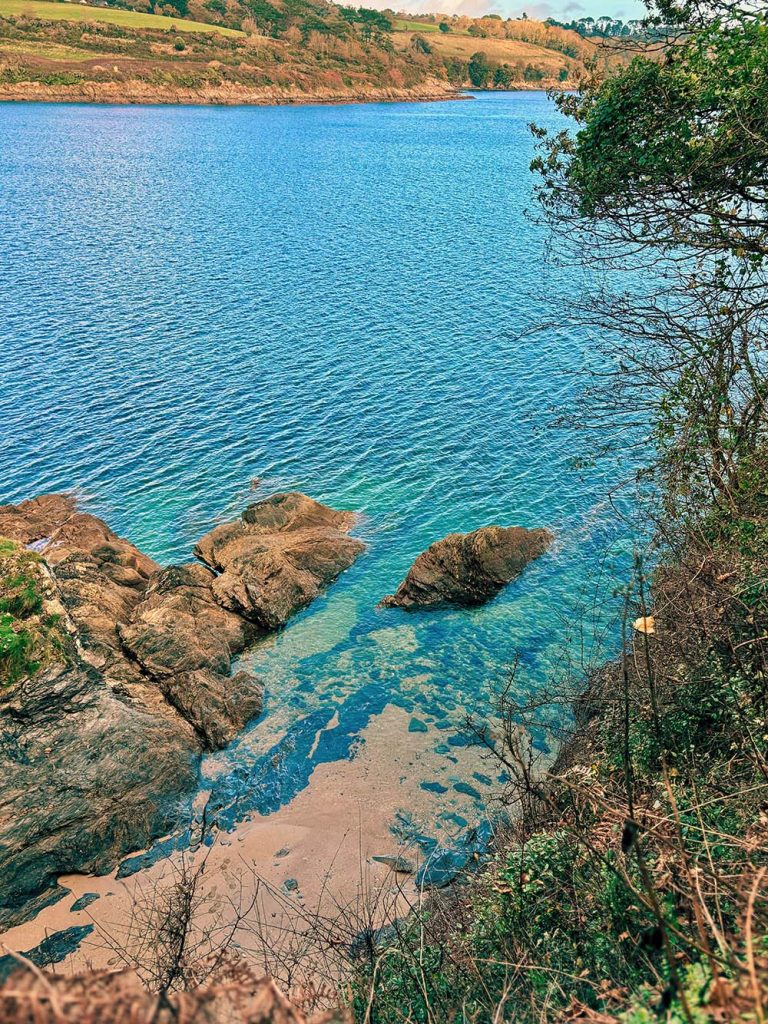 A scenic view of a clear blue body of water surrounded by rocks and greenery, with a gentle slope of land in the background.