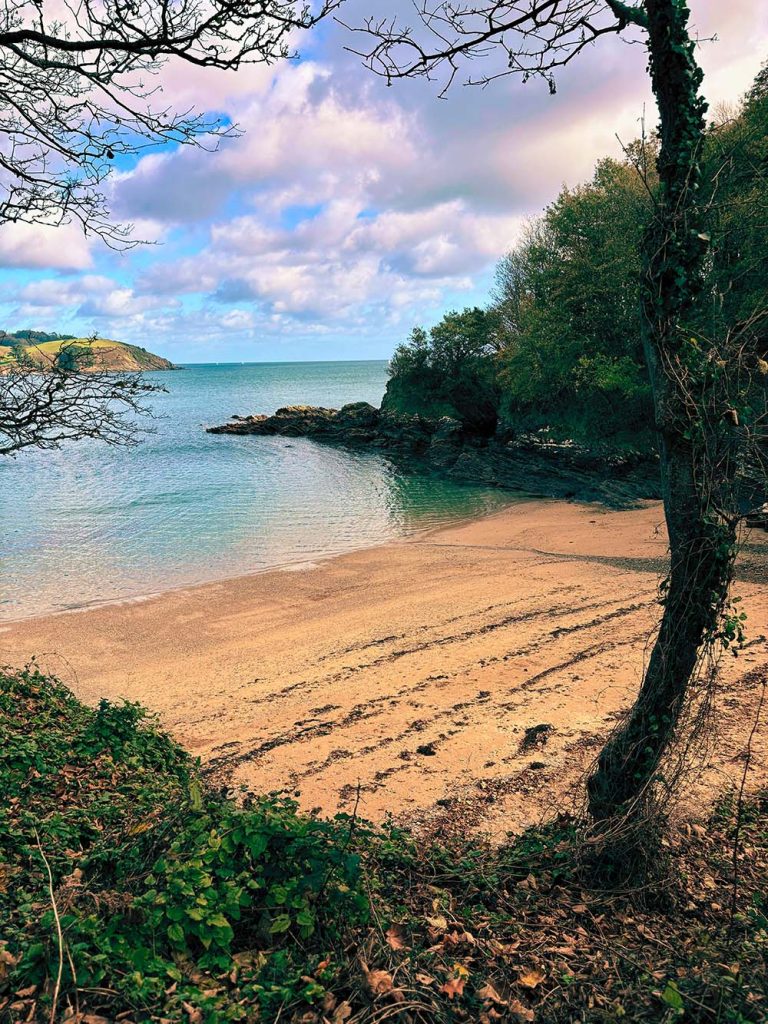 A serene beach scene with calm water, framed by trees and hills under a partly cloudy sky.