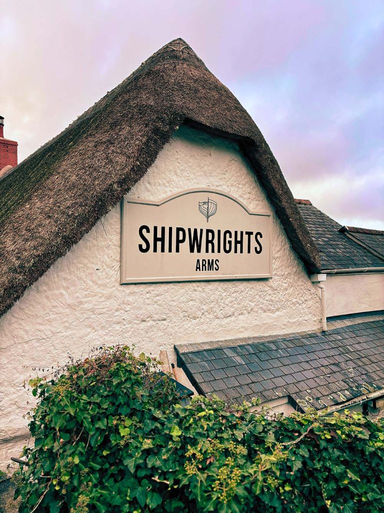 A thatched roof building with a sign that reads 'SHIPWRIGHTS ARMS' surrounded by greenery.