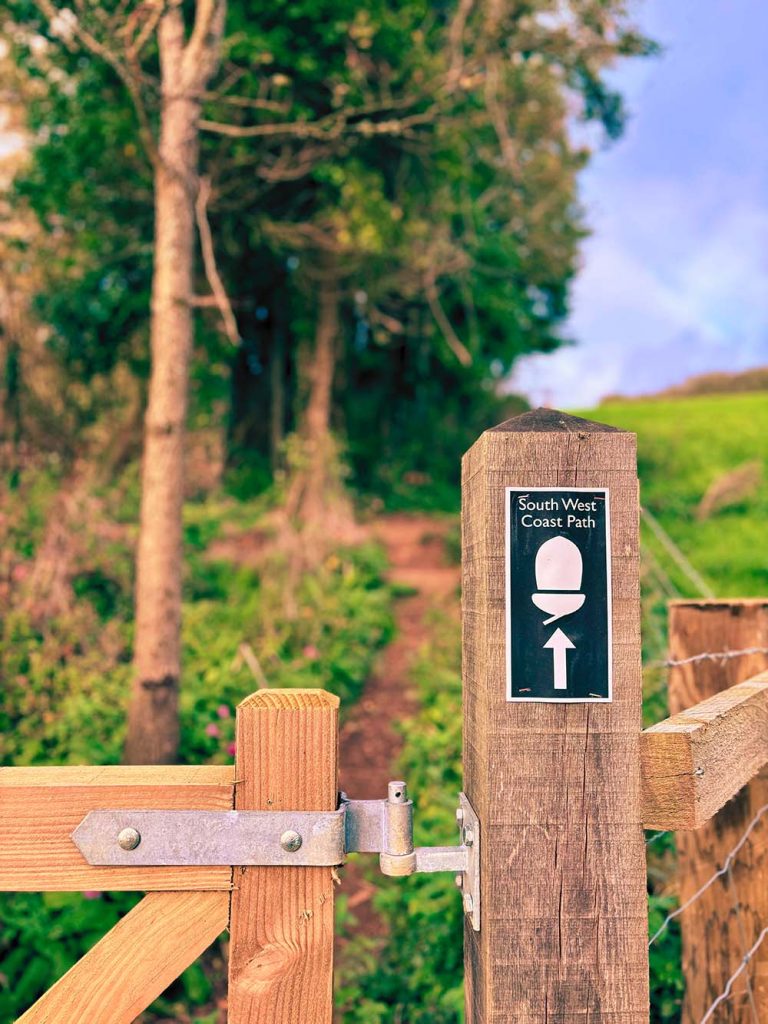 Signpost for the South West Coast Path next to a wooden gate, surrounded by trees and greenery.