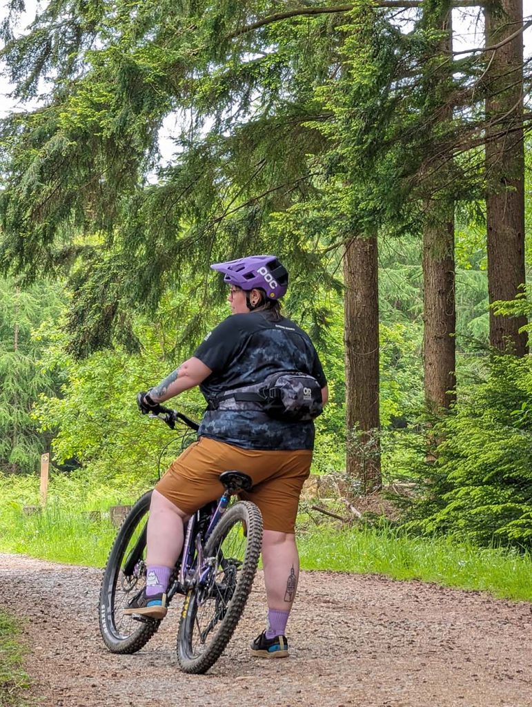 A person wearing a purple helmet and casual attire stands beside a mountain bike on a gravel path surrounded by lush green trees.