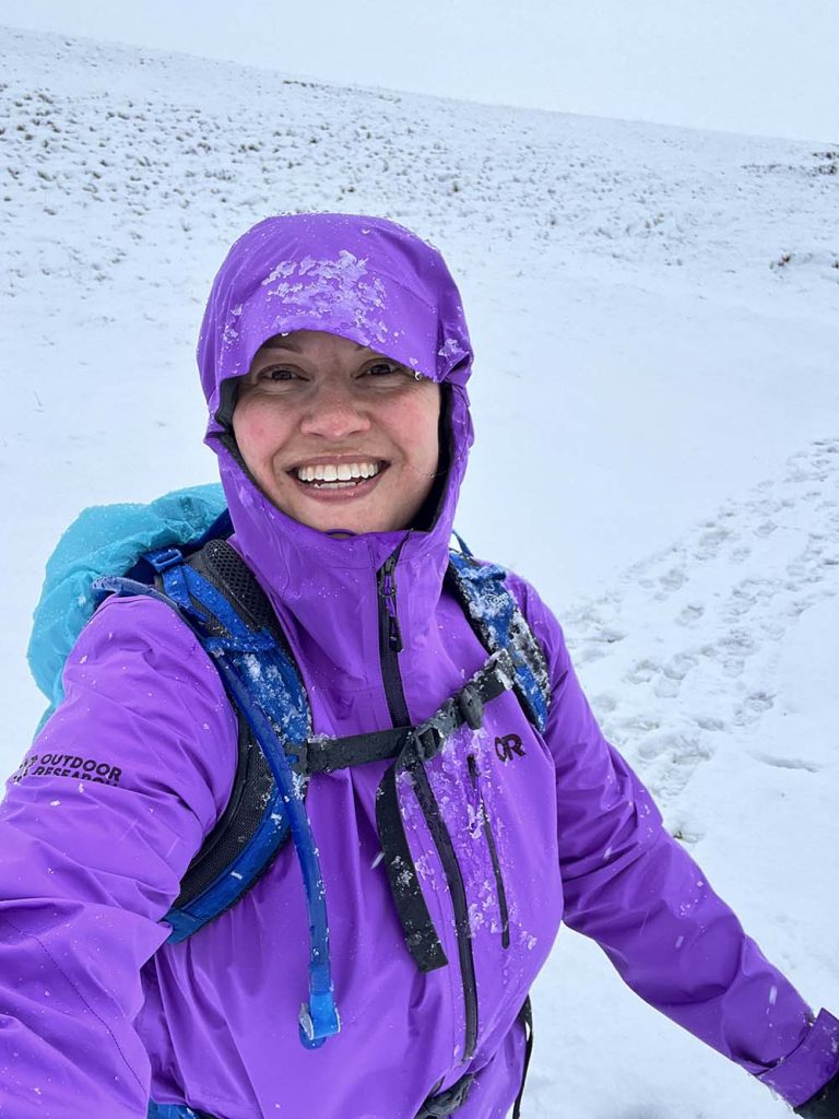 A smiling person wearing a purple waterproof jacket with snow on it, standing in a snowy landscape.
