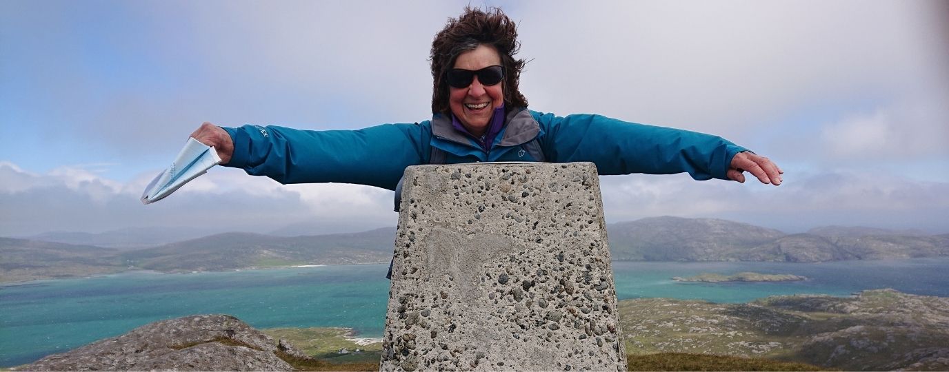 Sue Viccars with sunglasses joyfully leaning on a trig point at a scenic viewpoint, with a picturesque landscape of hills and a turquoise sea in the background.