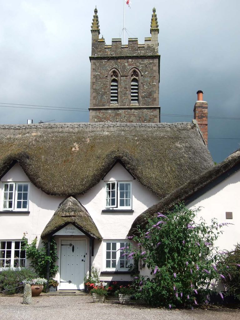 A traditional thatched cottage with a triangular roof and white walls, featuring a well-kept garden and a stone church tower in the background under a cloudy sky.