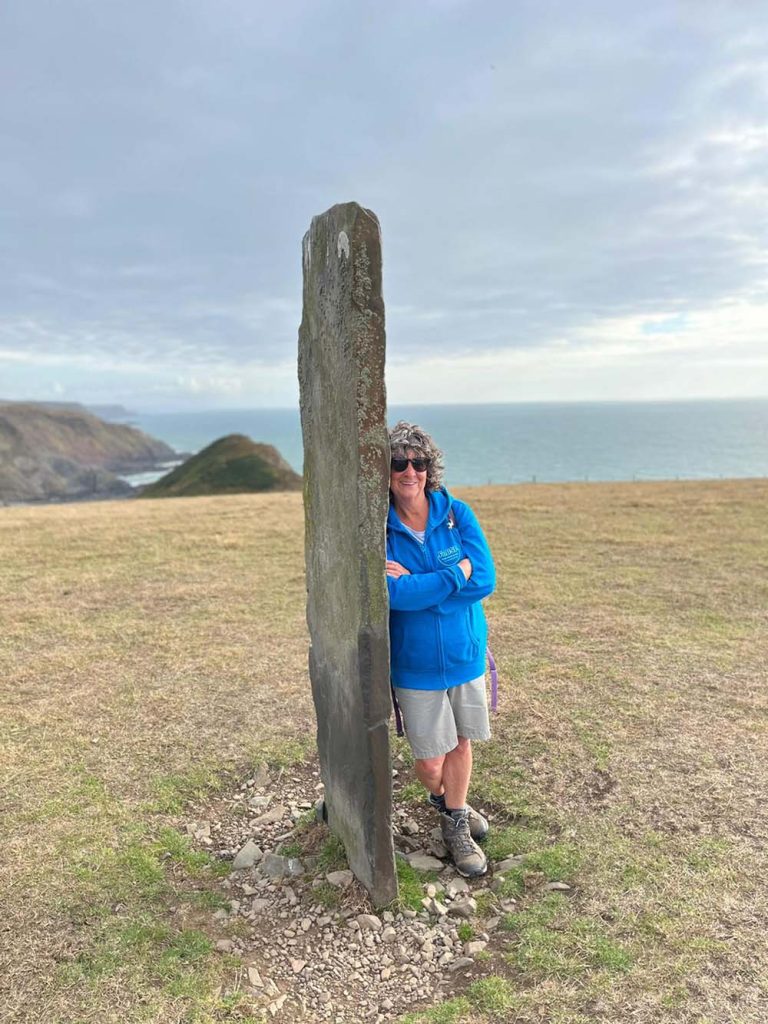 Person leaning against a tall stone in a scenic outdoor landscape with hills and ocean in the background.