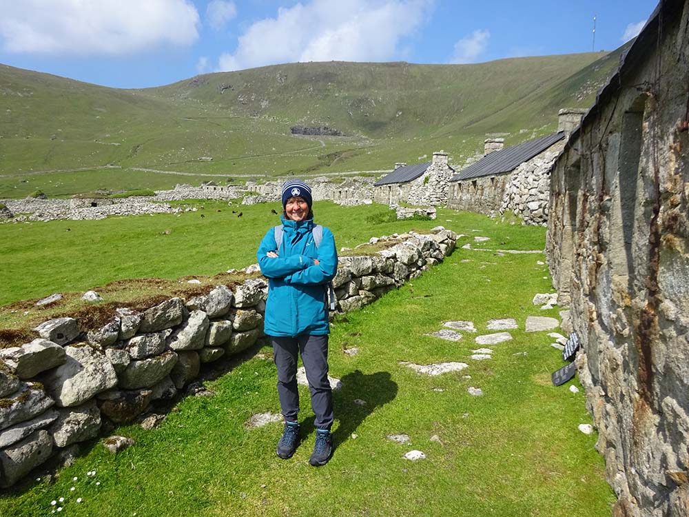 A person standing confidently in a green landscape beside a stone wall, with old structures and hills in the background on a sunny day.