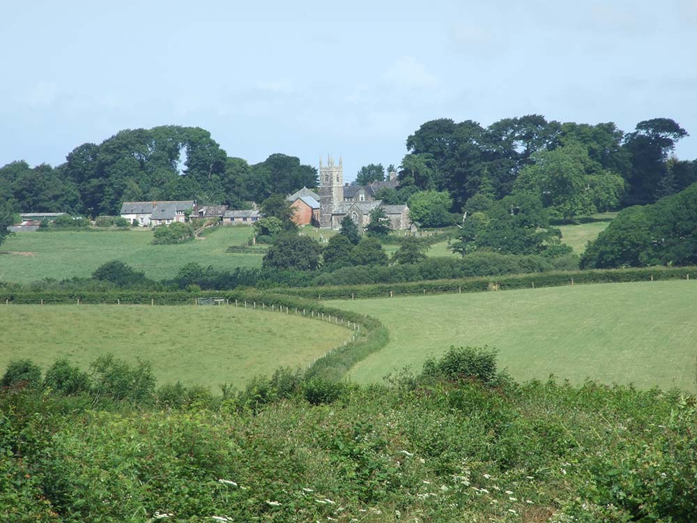 A scenic view of a rural countryside with a small village, featuring traditional houses and a church amidst lush green fields and trees.