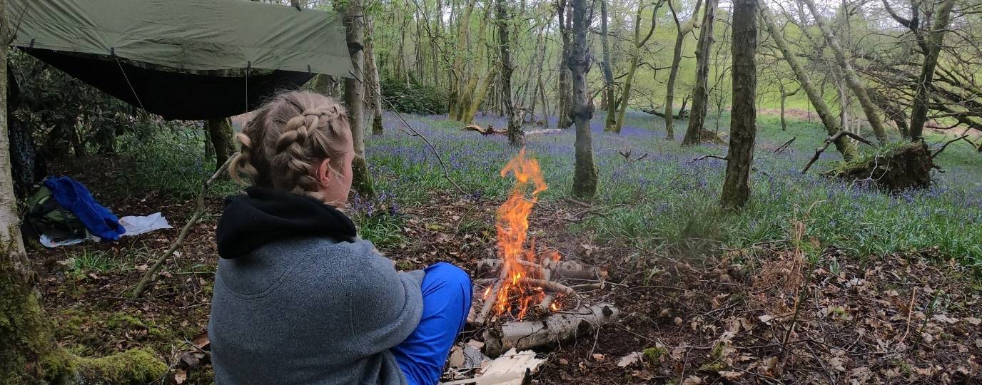a woman sat in front of the fire in a bluebell wood