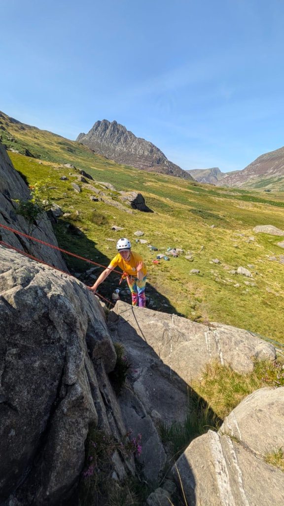 A woman waves while standing on a steep rock, wearing a harness and rope