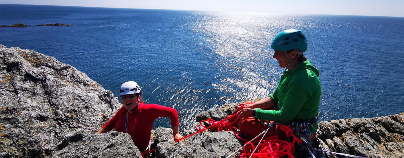 A woman climbs over a cliff top while partner holds a rope